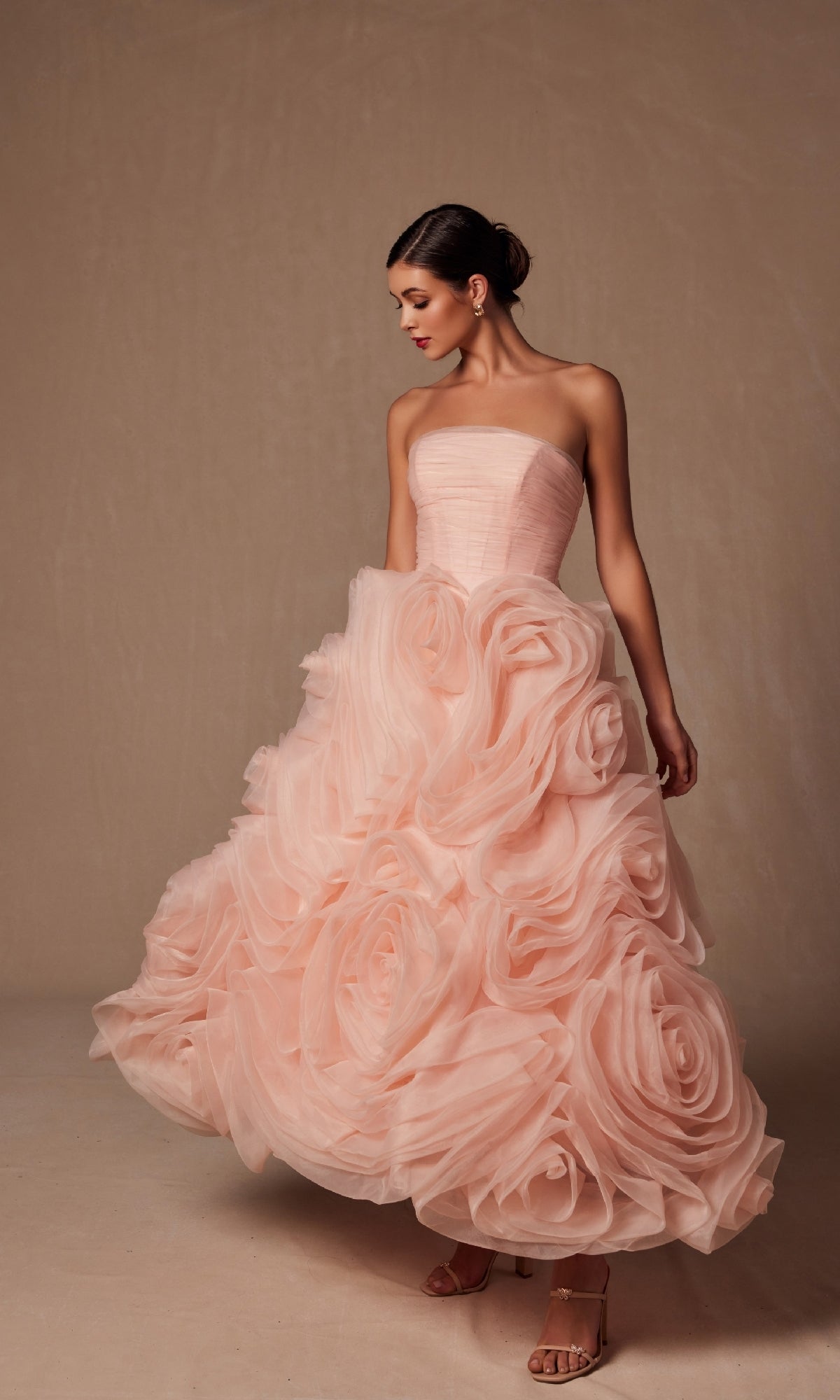 A woman stands gracefully against a plain beige background, wearing the Nox Anabel JQ1037 Strapless Peach Prom Ball Gown with an oversized rosette skirt and sculpted roses.